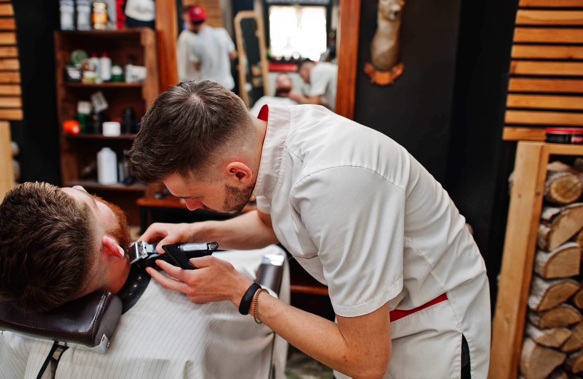 Young bearded man getting haircut by hairdresser while sitting in chair at barbershop. Barber soul.
