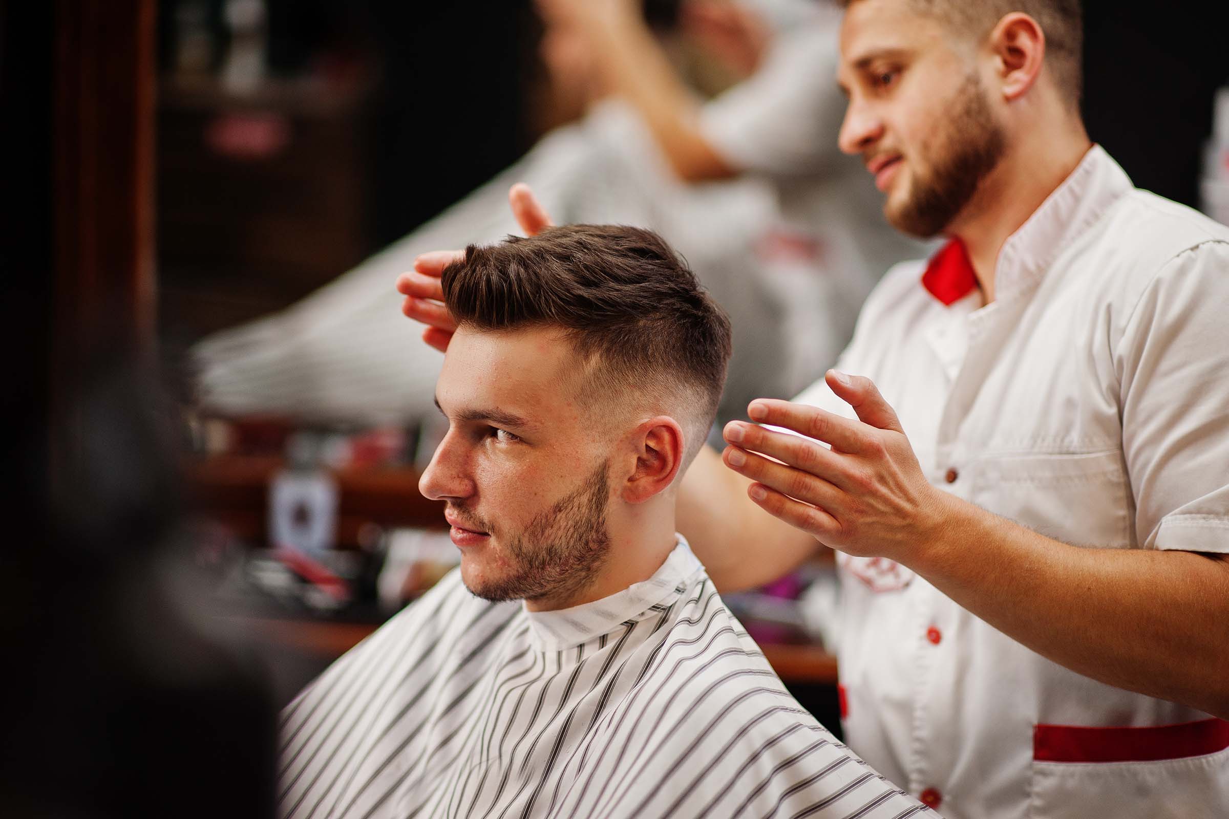 Young bearded man getting haircut by hairdresser while sitting in chair at barbershop. Barber soul.